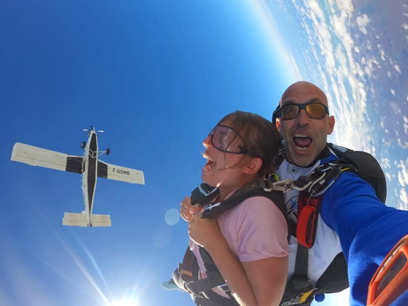 Une femme criant de joie en chute libre avec son instructeur, un avion blanc visible au loin dans un ciel bleu intense.