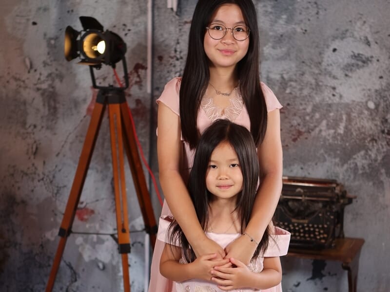 Deux sœurs en robes roses posant ensemble, l’aînée tenant la cadette dans ses bras, fond studio.
