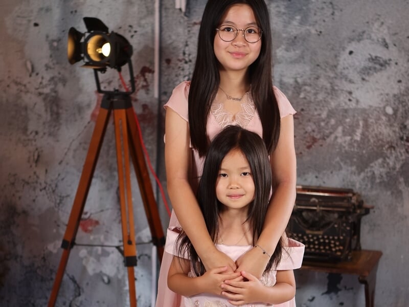 Deux sœurs en robes roses posant ensemble, l’aînée tenant la cadette dans ses bras, fond studio.