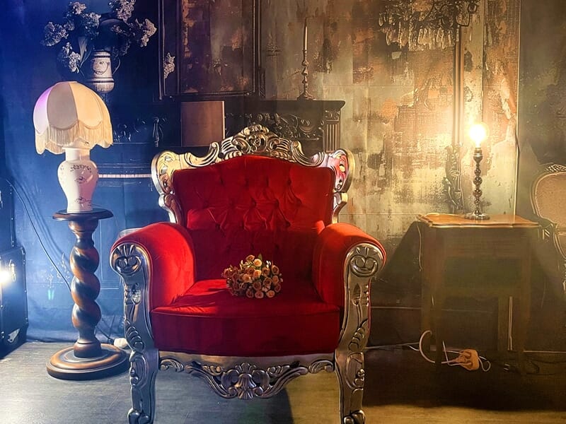 Fauteuil rouge capitonné au style baroque, entouré de lampadaires et de chandeliers dans un décor vintage.