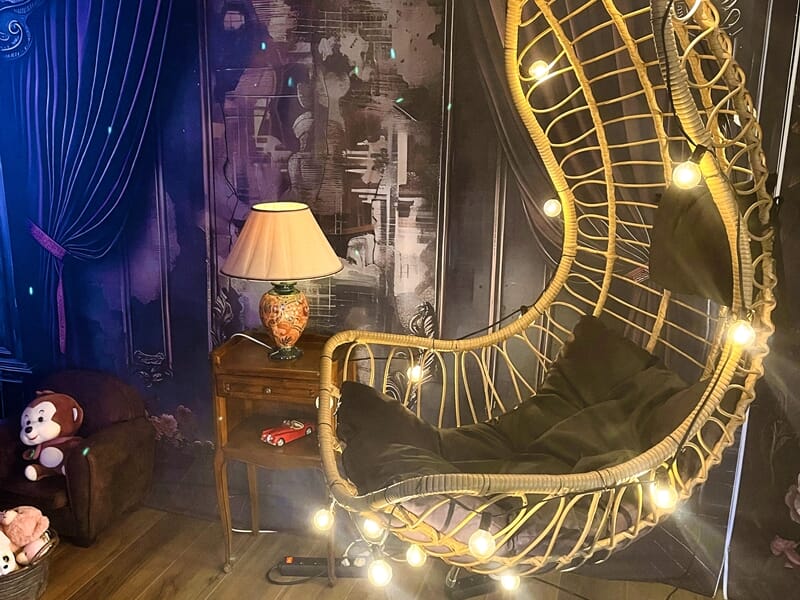 Chaise suspendue en rotin décorée de guirlandes lumineuses, avec coussin noir, dans un coin chaleureux au décor sombre et boisé.