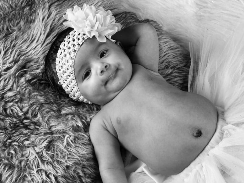 Bébé allongé sur un plaid poilu, bandeau à fleur, main derrière la tête, en noir et blanc.