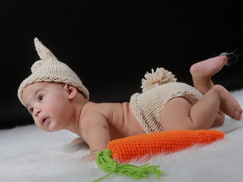 Bébé allongé sur un plaid blanc, bonnet et short en laine crème, tenant une carotte en tricot.