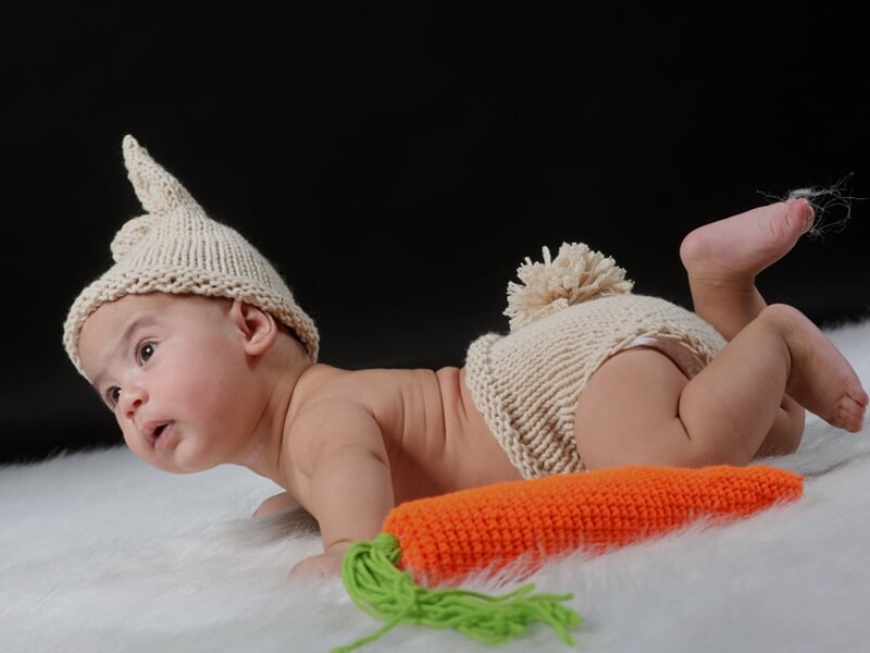 Bébé allongé sur un plaid blanc, bonnet et short en laine crème, tenant une carotte en tricot.