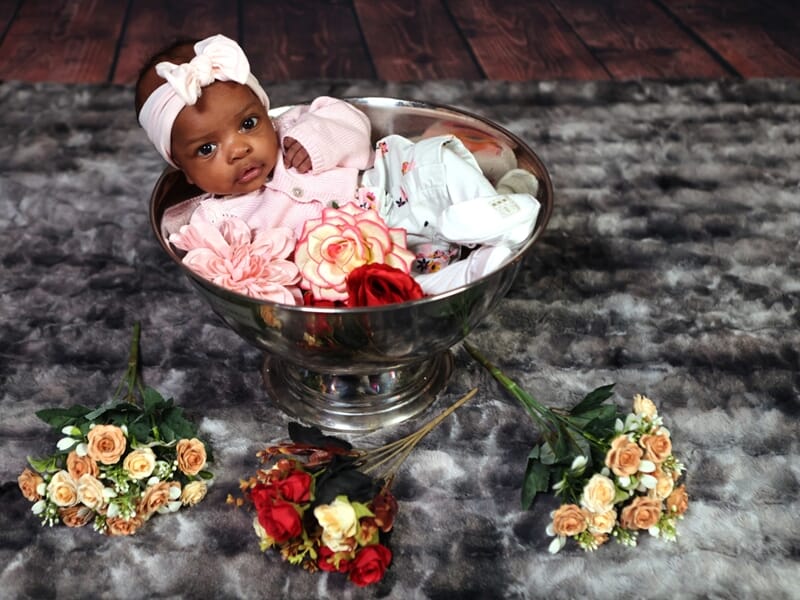 Bébé dans un grand saladier argenté avec fleurs, sur un tapis gris, bouquets autour.