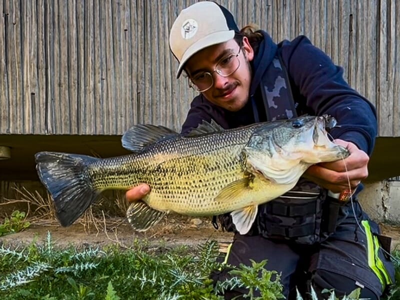 Pêcheur accroupi tenant un black-bass aux reflets dorés, pêché près d’un bâtiment en bois.