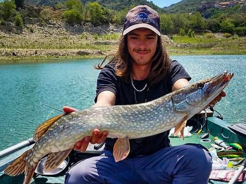 Homme assis sur une barque, présentant fièrement un brochet allongé.