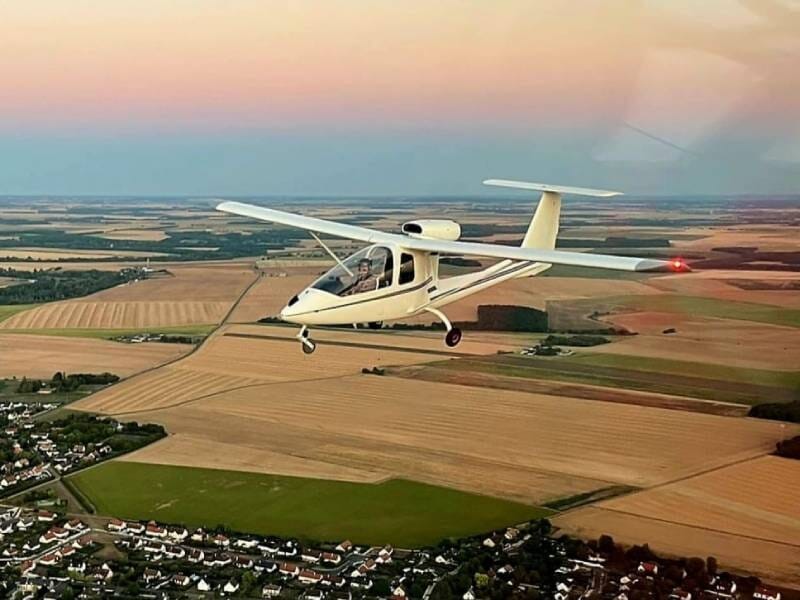Un avion ULM Sky Arrow 500 FT en vol au-dessus des champs avec un ciel bleu-rose en fond.