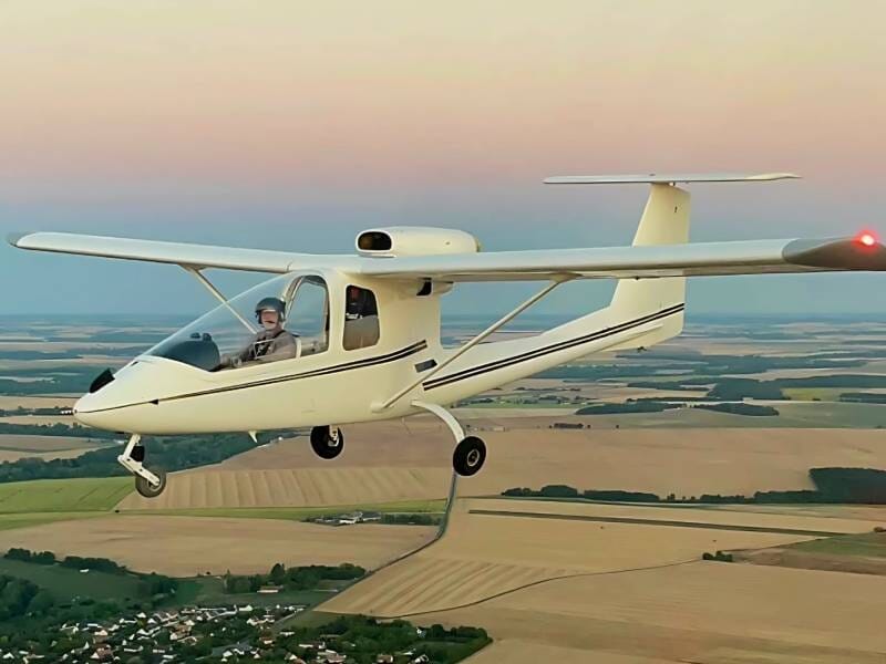 Un avion ULM Sky Arrow 500 FT en vol au-dessus des champs avec un ciel bleu-rose en fond.
