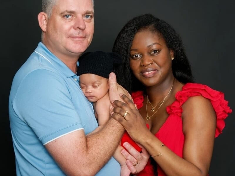 Couple tenant leur nouveau-né endormi, l’homme en polo bleu et la femme en robe rouge souriante, sur fond noir.