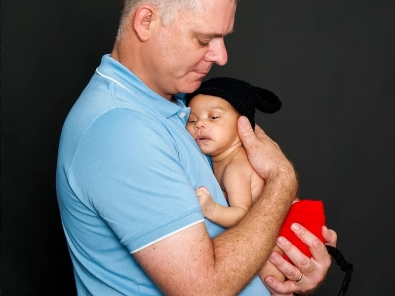 Homme en polo bleu tenant tendrement un nouveau-né endormi portant un bonnet noir et une couverture rouge.