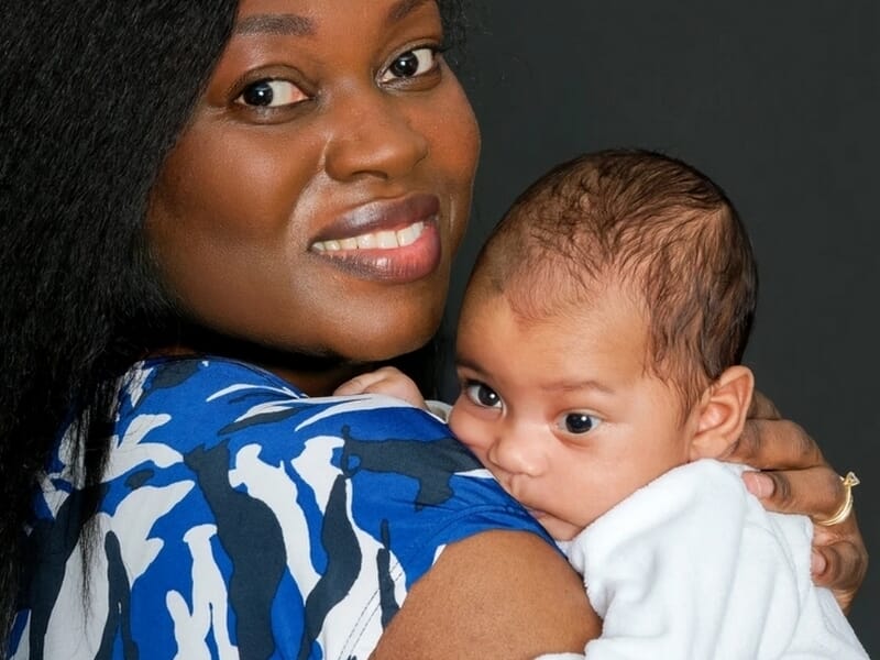 Femme souriante en robe bleue et blanche portant un bébé éveillé contre son épaule, sur fond noir.