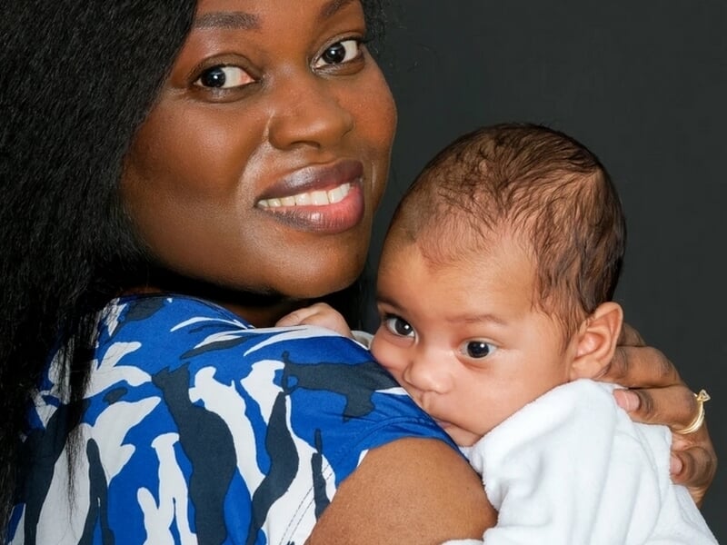 Femme souriante en robe bleue et blanche portant un bébé éveillé contre son épaule, sur fond noir.