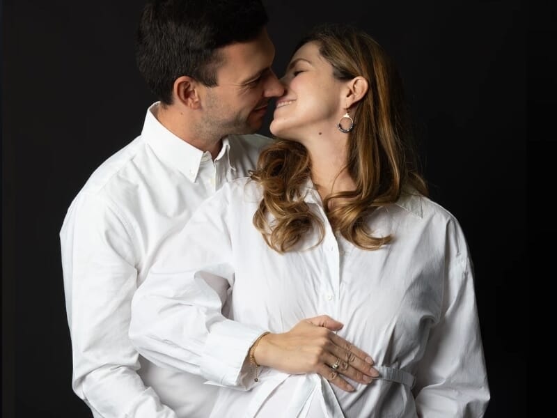 Couple s’embrassant tendrement, la femme enceinte en chemise blanche tenant son ventre, sur fond noir.