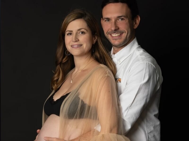 Couple souriant, la femme enceinte en robe beige transparente et brassière noire, l’homme en chemise blanche.