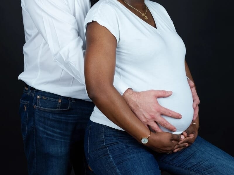 Mains d’un couple posées sur le ventre arrondi de la femme enceinte, tous deux vêtus de jeans et hauts blancs.