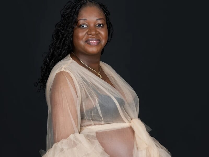 Femme enceinte souriante portant une robe en tulle beige translucide, posant de profil sur fond noir.