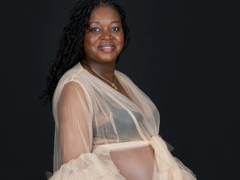 Femme enceinte souriante portant une robe en tulle beige translucide, posant de profil sur fond noir.