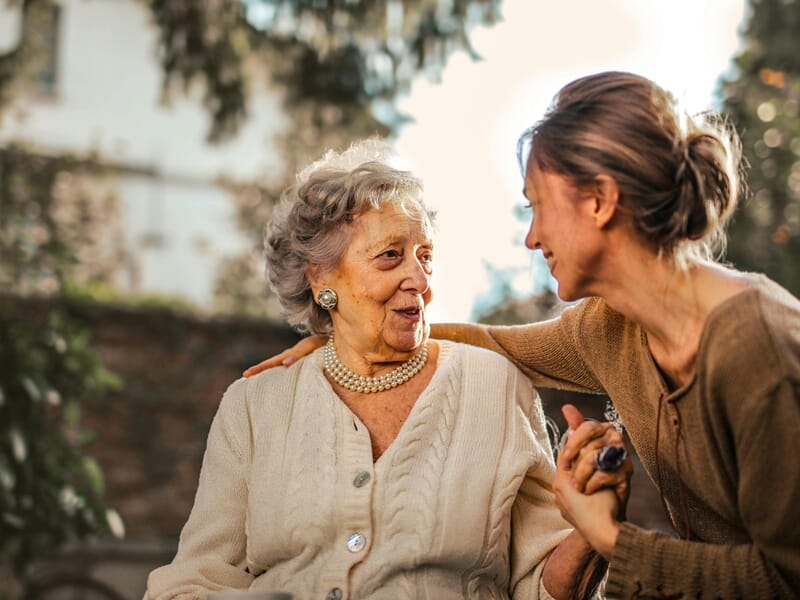 Femme âgée souriante discutant avec une femme plus jeune, assises dehors dans un jardin.