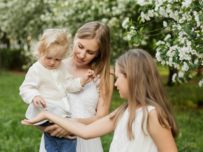 Mère et ses deux filles observant la main de l’aînée dans un jardin fleuri au printemps.