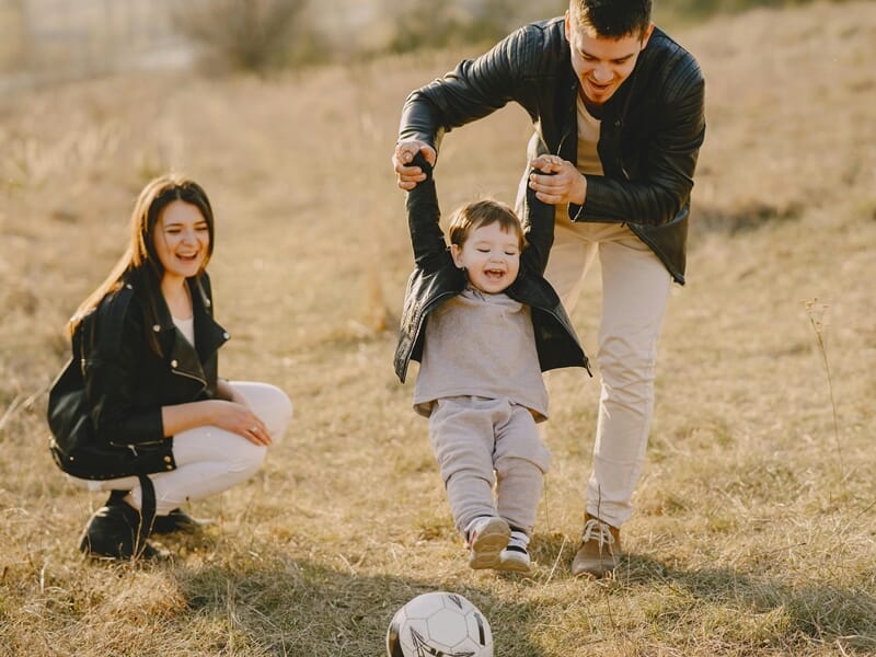 Parents jouant avec leur enfant sur un terrain, le soulevant par les bras près d’un ballon.