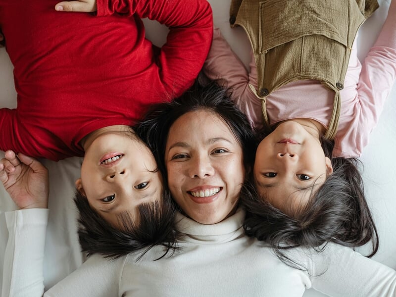 Mère allongée sur un lit avec ses deux enfants, têtes rapprochées et sourires complices.