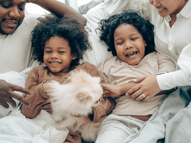 Famille souriante allongée sur un lit avec deux enfants jouant avec un petit chien blanc.