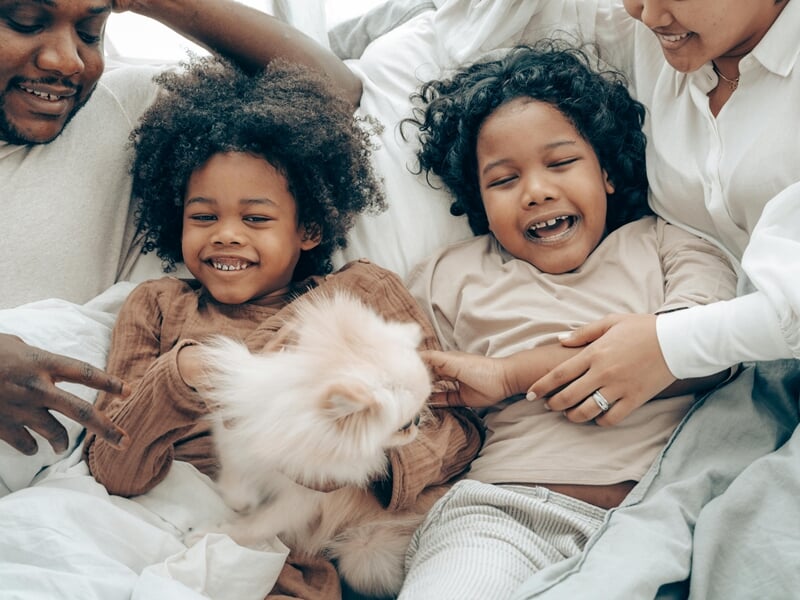 Famille souriante allongée sur un lit avec deux enfants jouant avec un petit chien blanc.