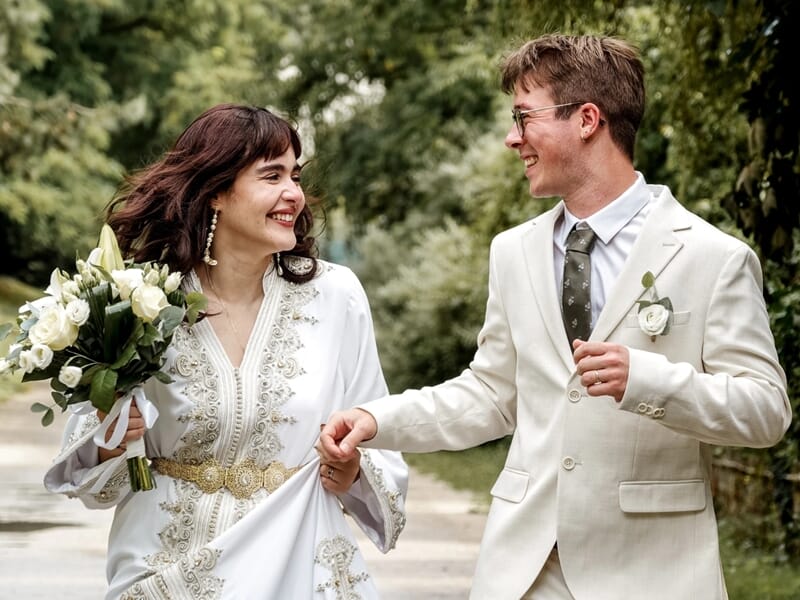 Jeunes mariés marchant main dans la main, la mariée tenant un bouquet de fleurs blanches.