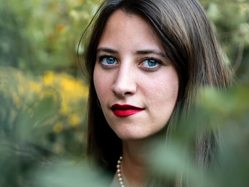 Portrait d'une femme aux yeux bleus et lèvres rouges, entourée de feuillage vert et jaune.