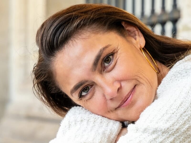 Femme souriante appuyée sur ses bras, portant un pull blanc et de grandes boucles d'oreilles dorées.