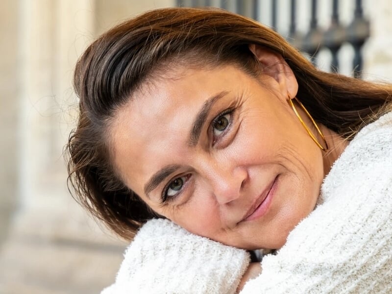 Femme souriante appuyée sur ses bras, portant un pull blanc et de grandes boucles d'oreilles dorées.