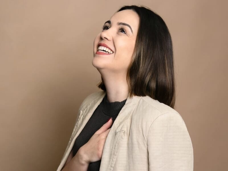 Femme souriante regardant vers le haut, main sur la poitrine, vêtue d'un blazer beige et haut noir.