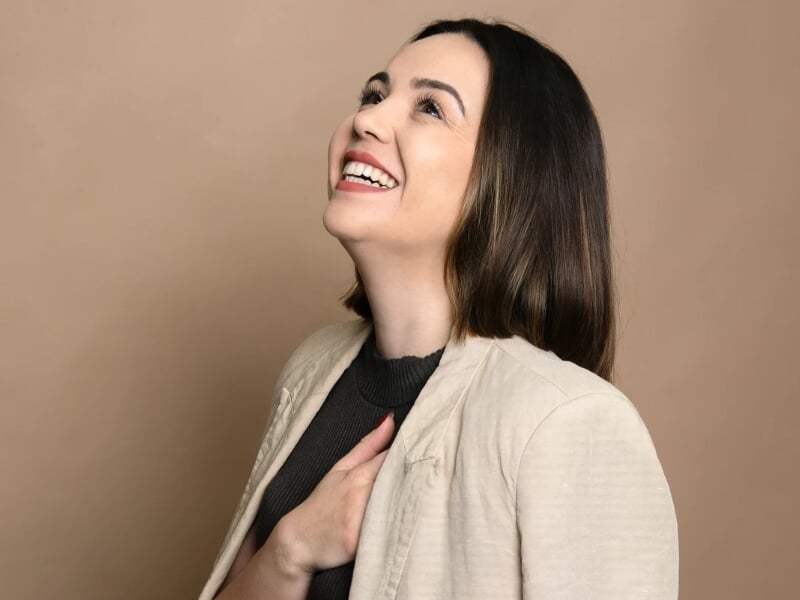 Femme souriante regardant vers le haut, main sur la poitrine, vêtue d'un blazer beige et haut noir.