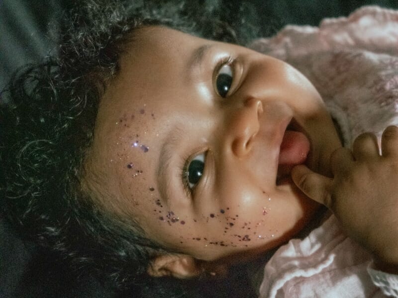 Bébé allongé sur le côté, le visage couvert de paillettes scintillantes, souriant et portant la main à sa bouche.