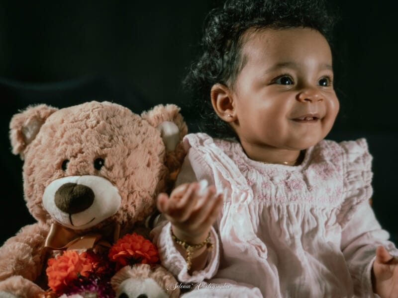 Bébé souriant en robe claire assis à côté d’un gros ours en peluche beige et de fleurs colorées