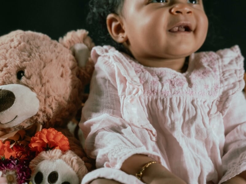 Portrait d’un bébé souriant en robe claire, assis à côté d’un ours en peluche et de fleurs.