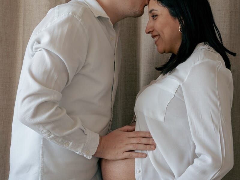 Couple en séance photo, l’homme embrasse le front de sa compagne enceinte tout en posant sa main sur son ventre.