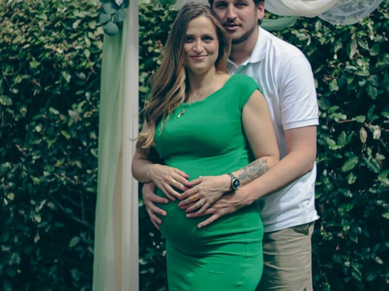 Couple en extérieur, la femme enceinte en robe verte pose devant une arche décorée de voiles et de fleurs.