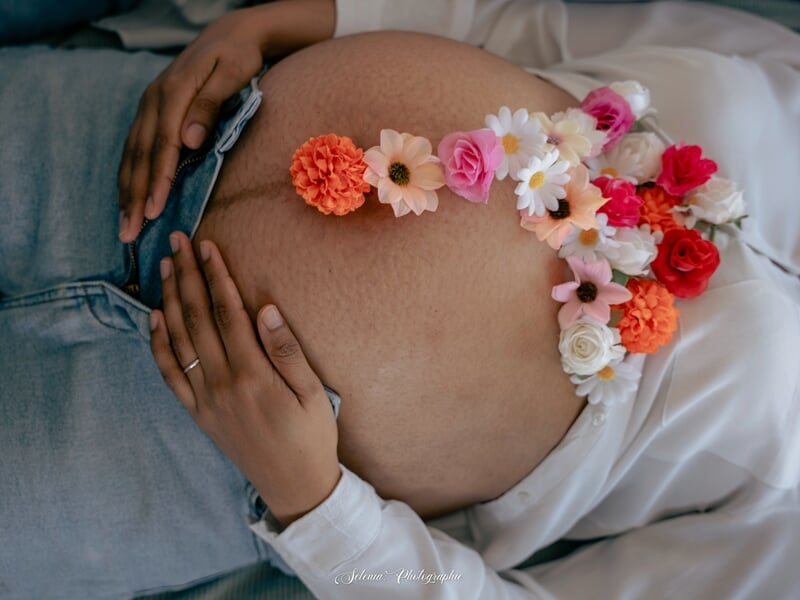 Femme enceinte allongée avec un jean entrouvert, son ventre orné de fleurs colorées.