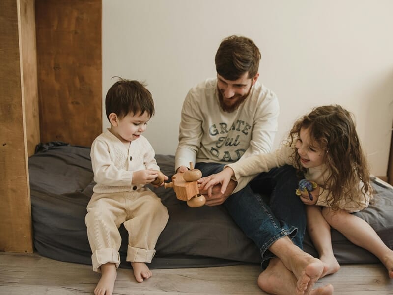 Père jouant avec ses deux enfants assis par terre, partageant un moment complice avec un jouet en bois.