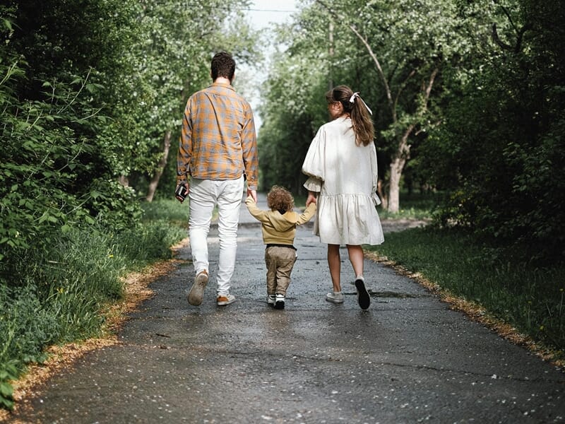 Couple de dos tenant la main de leur enfant en bas âge, marchant ensemble sur un chemin bordé d’arbres.