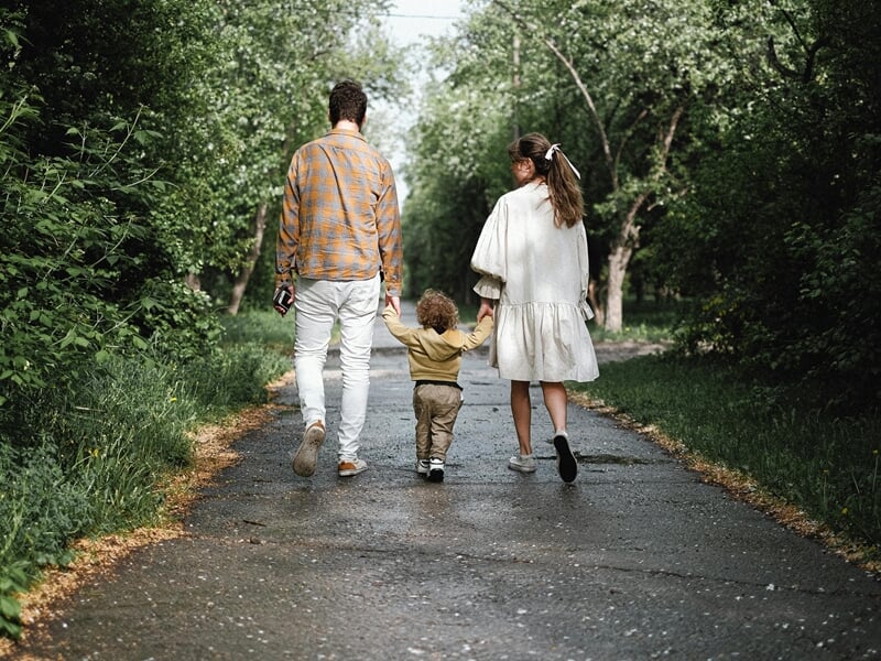 Couple de dos tenant la main de leur enfant en bas âge, marchant ensemble sur un chemin bordé d’arbres.