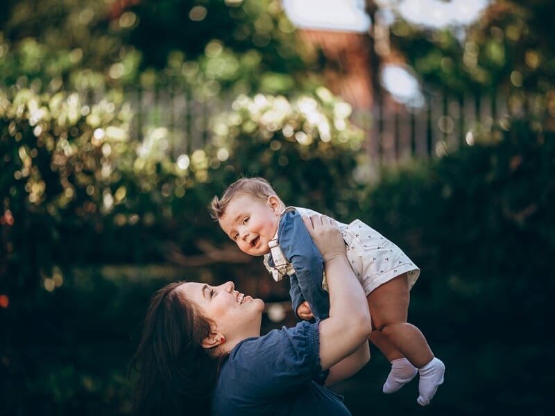 Mère levant son bébé en riant dans un jardin, moments de tendresse et de complicité.