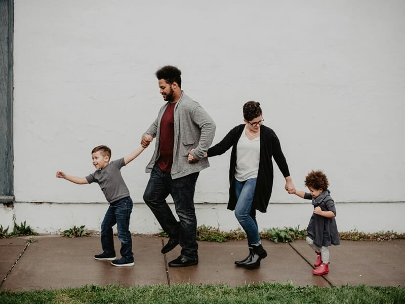 Famille marchant main dans la main sur un trottoir, deux enfants tenus par leurs parents.