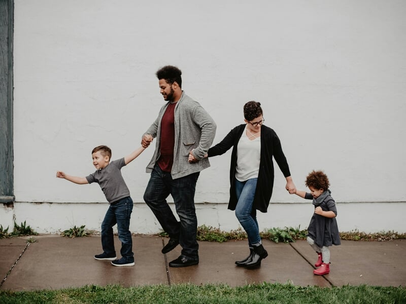 Famille marchant main dans la main sur un trottoir, deux enfants tenus par leurs parents.