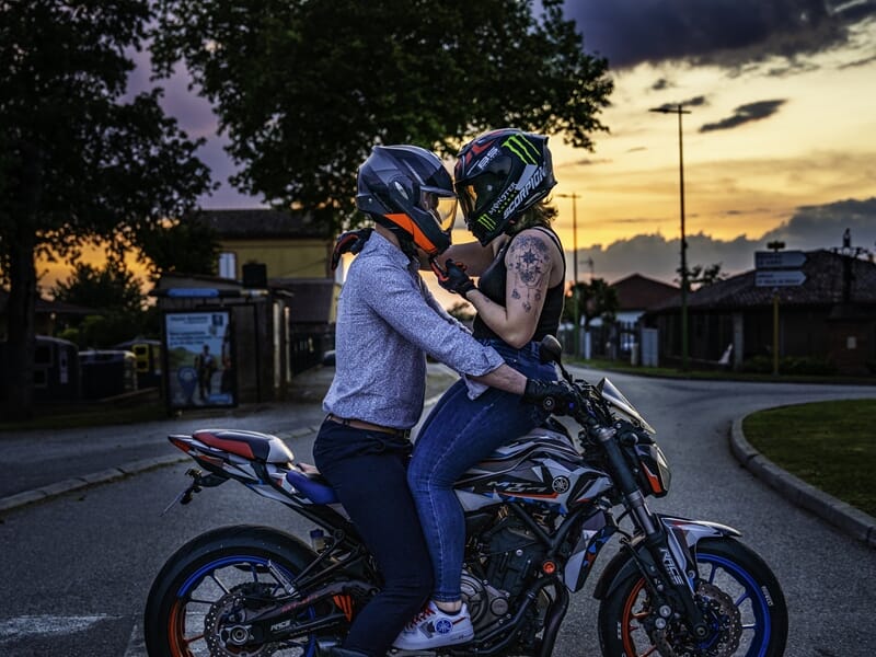 Couple de motards casqués enlacés sur une moto au coucher du soleil.