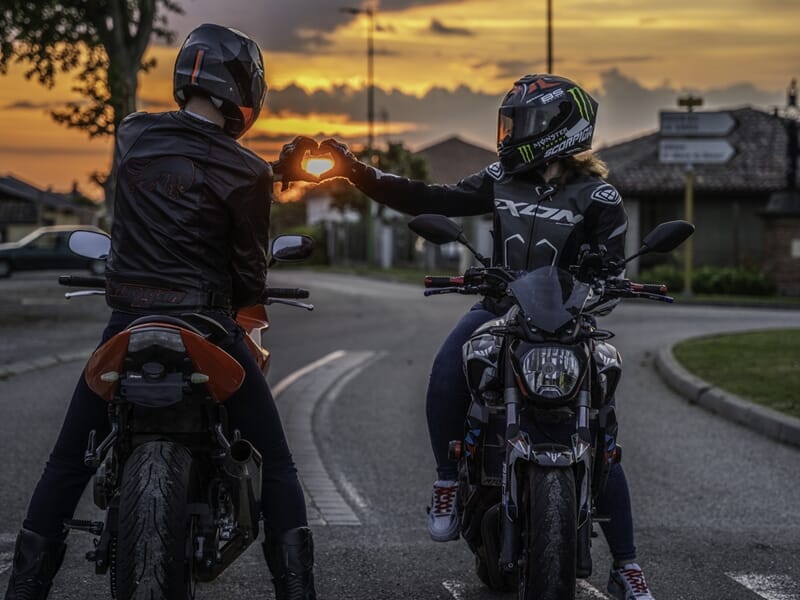 Deux motards formant un cœur avec leurs mains face au coucher de soleil.