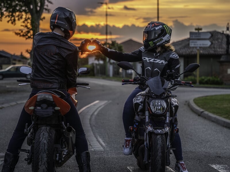 Deux motards formant un cœur avec leurs mains face au coucher de soleil.