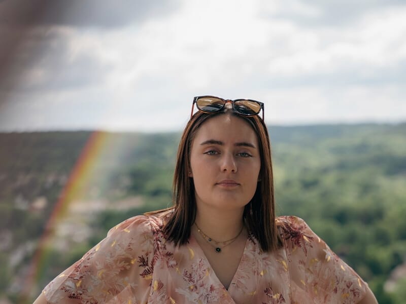 Jeune femme en robe fleurie avec ceinture noire, posant devant un panorama vallonné, un léger arc-en-ciel visible à gauche.
